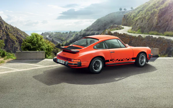 An orange Porsche 911 Carrera is parked on a winding road, surrounded by scenic hills, showcasing its sleek design against a stunning landscape backdrop.