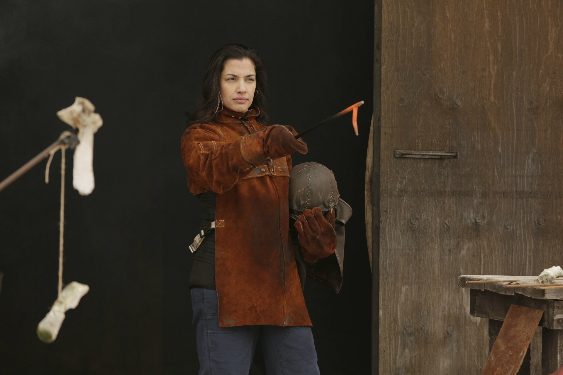 Wynonna Earp TV show: woman in a brown leather coat holds a helmet and knife beside a rustic door — 2K Quad HD PC desktop wallpaper and background.