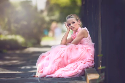 A blonde little girl in a flowing pink dress sits thoughtfully against a dark fence, captured with depth of field in a vibrant 4K Ultra HD photography background.