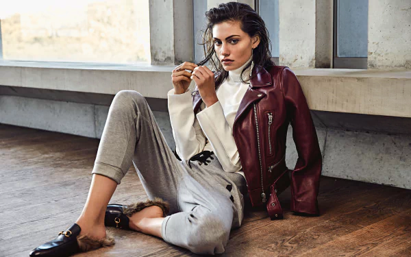 Brunette woman with brown eyes in a burgundy leather jacket and neutral trousers, seated by a large industrial window — HD PC desktop wallpaper.