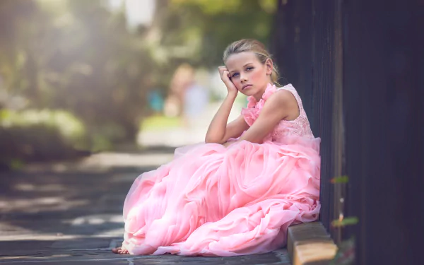 A blonde little girl in a flowing pink dress sits thoughtfully against a dark fence, captured with depth of field in a vibrant 4K Ultra HD photography background.