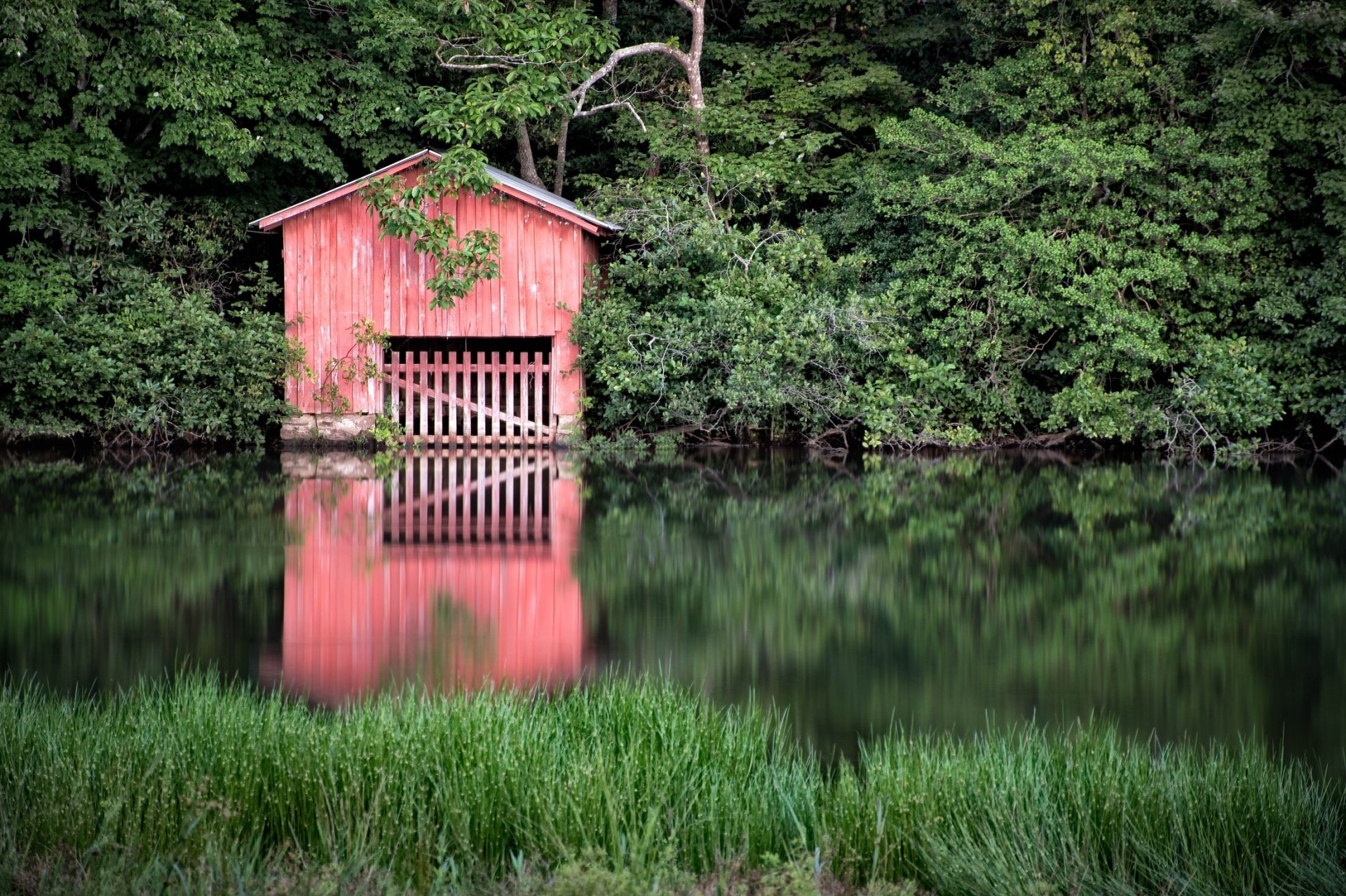 Red man-made boathouse on a riverbank under dense trees, its façade and surrounding foliage reflected in calm water — HD PC desktop wallpaper/background.