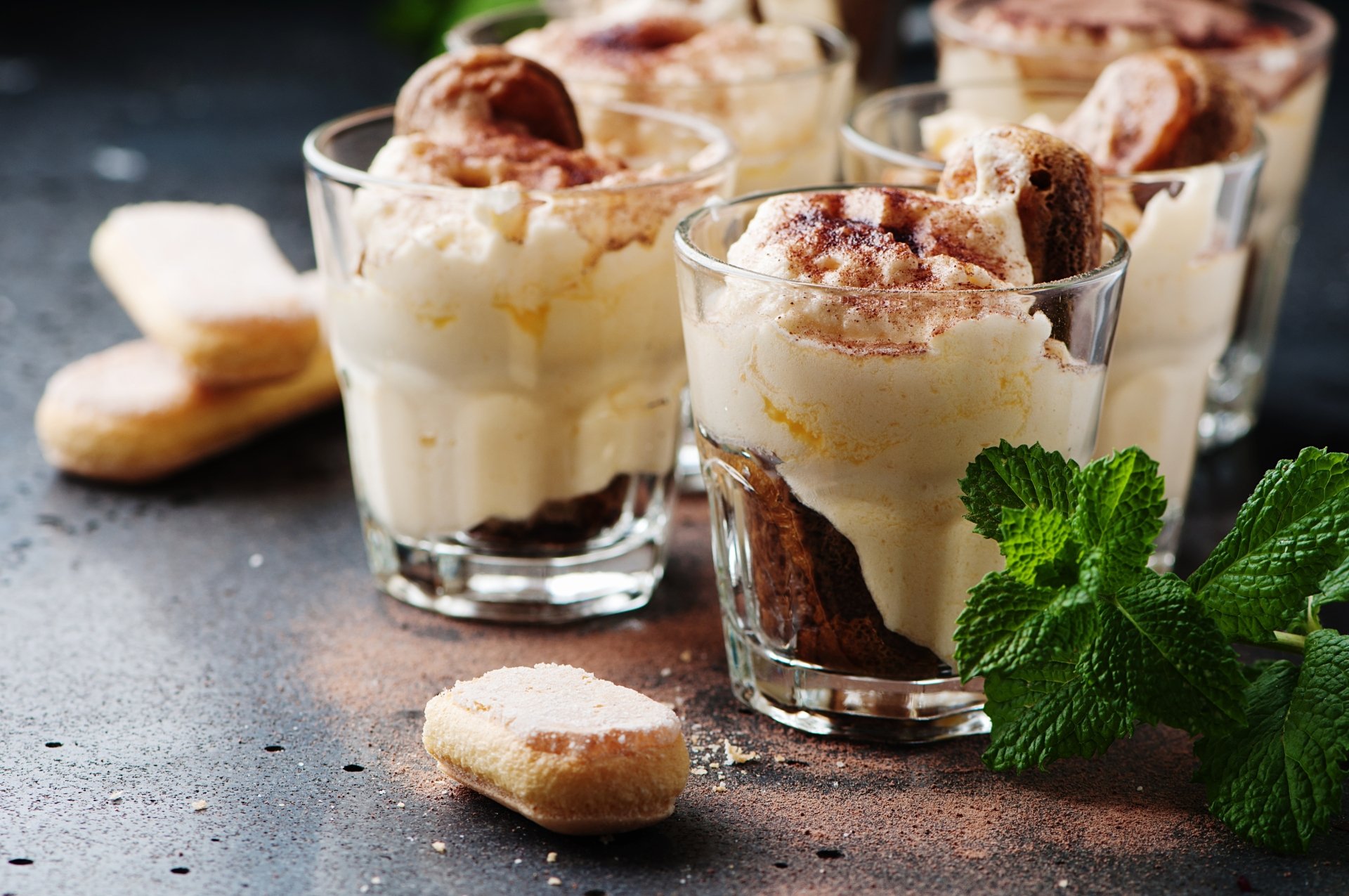 Creamy dessert served in clear glass cups with layers of biscuit, garnished with cocoa powder and fresh mint, captured in 4K Ultra HD for a PC desktop wallpaper.