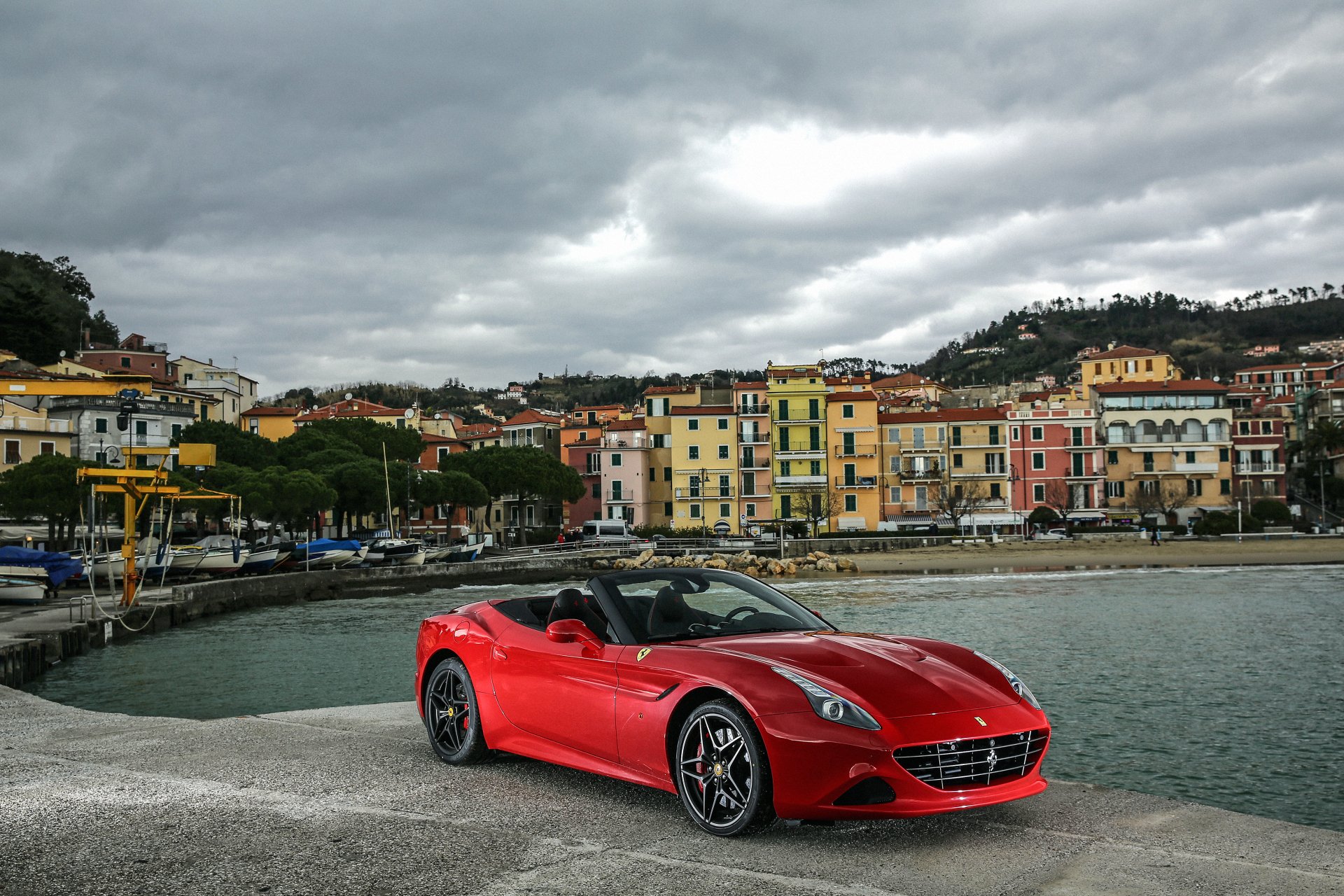 Red Ferrari California grand tourer parked on a seaside quay with colorful coastal buildings under a cloudy sky — 4K Ultra HD PC desktop wallpaper/background.