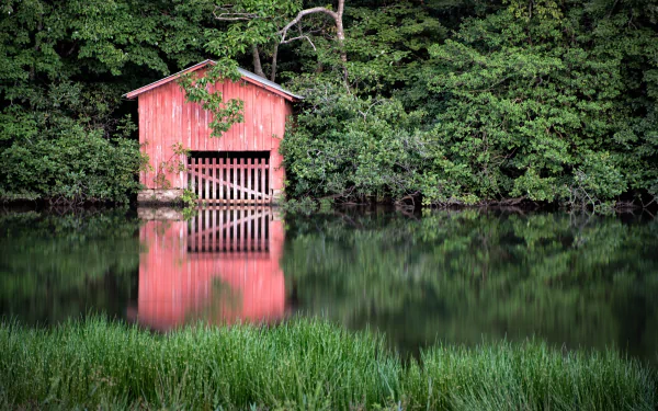 Red man-made boathouse on a riverbank under dense trees, its façade and surrounding foliage reflected in calm water — HD PC desktop wallpaper/background.