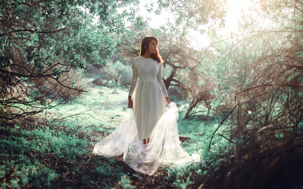 A brunette woman in a flowing white dress stands in a vibrant outdoor setting, surrounded by lush greenery and softly blurred sunlight, capturing a serene mood.