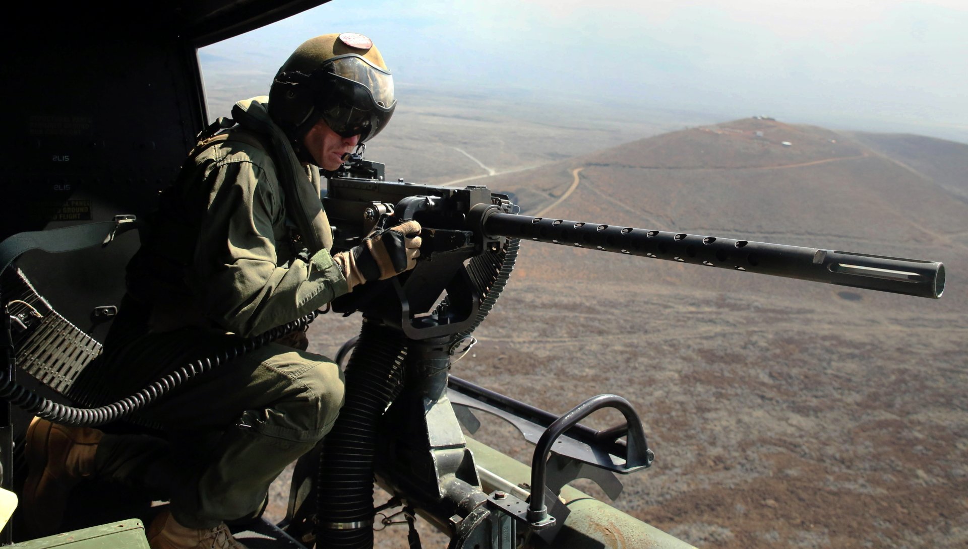A soldier in military gear aims a machine gun from a helicopter, overlooking a rugged landscape, captured in vibrant 4K Ultra HD detail.