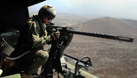 A soldier in military gear aims a machine gun from a helicopter, overlooking a rugged landscape, captured in vibrant 4K Ultra HD detail.