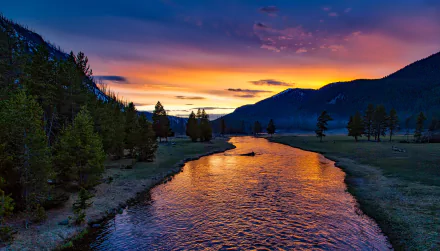 A scenic view of a river reflecting a vibrant sunset in Yellowstone National Park, framed by mountains and trees, capturing the serene beauty of wilderness at twilight.