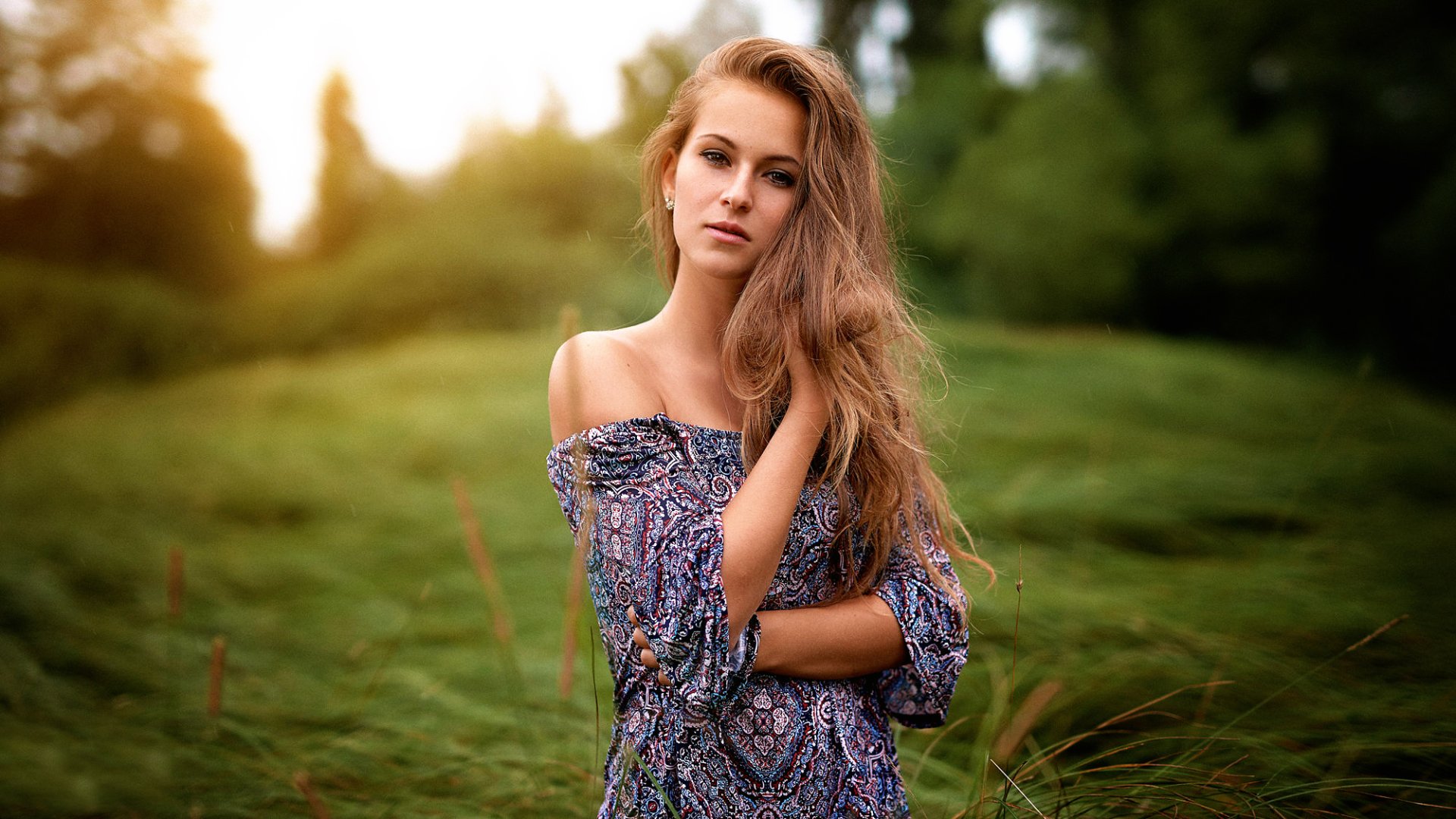 A blonde woman with brown eyes poses outdoors in a patterned top, surrounded by lush greenery, creating a serene and captivating HD desktop wallpaper.