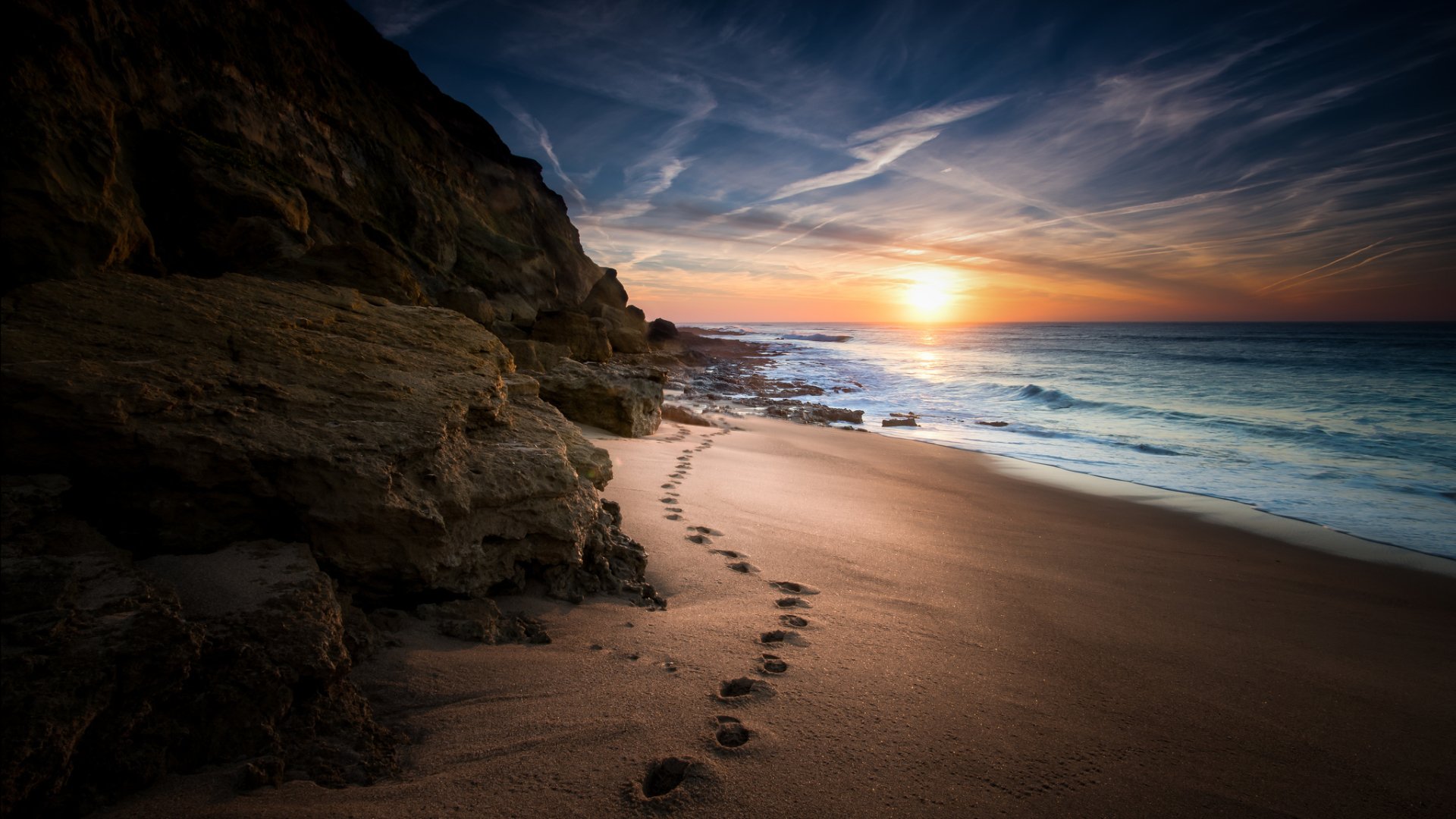 A serene beach scene at sunset, featuring footprints in the sand leading towards the horizon over the ocean, framed by rocky cliffs, capturing the beauty of nature.