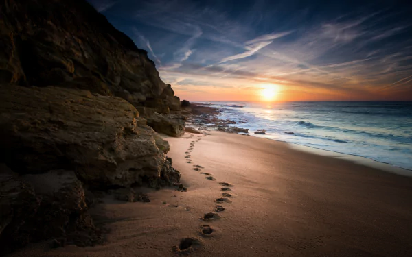 A serene beach scene at sunset, featuring footprints in the sand leading towards the horizon over the ocean, framed by rocky cliffs, capturing the beauty of nature.