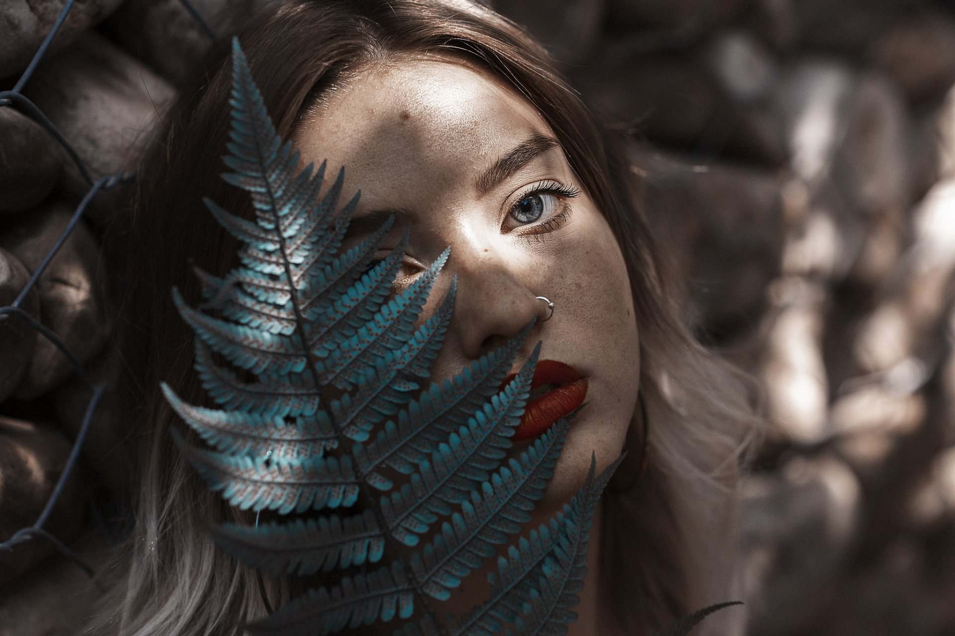 HD desktop wallpaper of a woman with blue eyes and red lipstick partially obscured by a fern, her face softly lit with natural shadows.