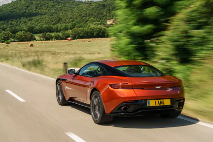 An orange Aston Martin DB11 grand tourer is seen speeding along a scenic road surrounded by lush greenery, captured in stunning 4K Ultra HD.