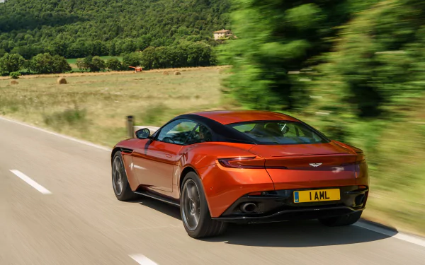 An orange Aston Martin DB11 grand tourer is seen speeding along a scenic road surrounded by lush greenery, captured in stunning 4K Ultra HD.
