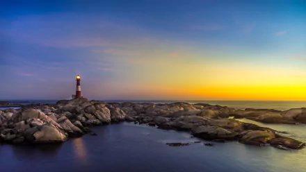 A serene evening landscape featuring a lighthouse standing on rocky shores under a vibrant horizon, perfect as a high-definition desktop wallpaper or background.