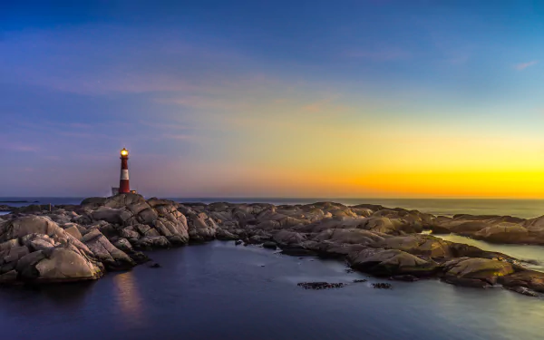 A serene evening landscape featuring a lighthouse standing on rocky shores under a vibrant horizon, perfect as a high-definition desktop wallpaper or background.