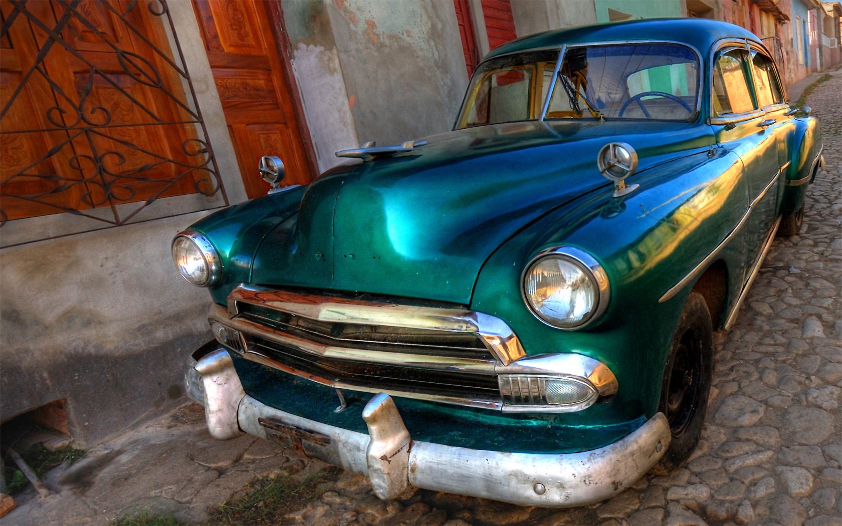 A classic blue-green vintage car parked on a cobblestone street, highlighting its charming details against a backdrop of rustic buildings. An HD desktop wallpaper capturing timeless beauty.