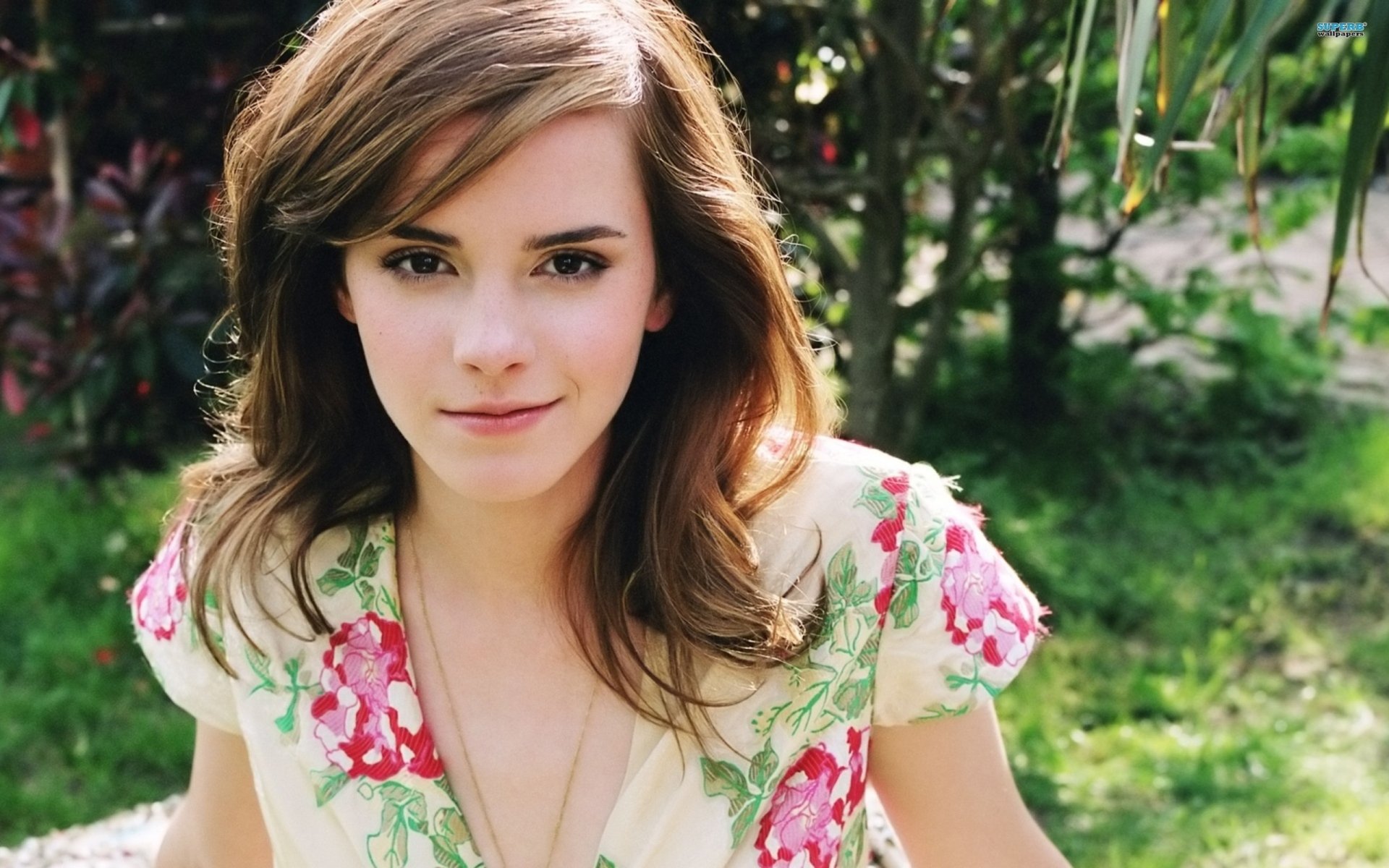 HD desktop wallpaper of actress Emma Watson, smiling outdoors in a floral dress, surrounded by lush greenery.