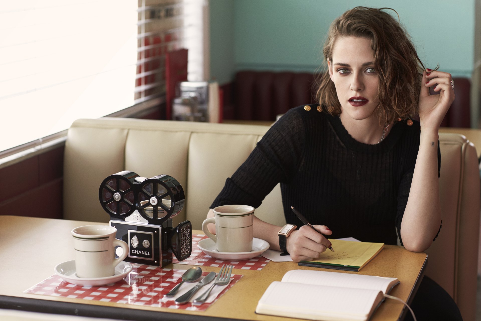 Kristen Stewart poses thoughtfully in a retro diner, with coffee cups and a vintage camera on the table, showcasing her stylish look in this 4K Ultra HD desktop wallpaper.