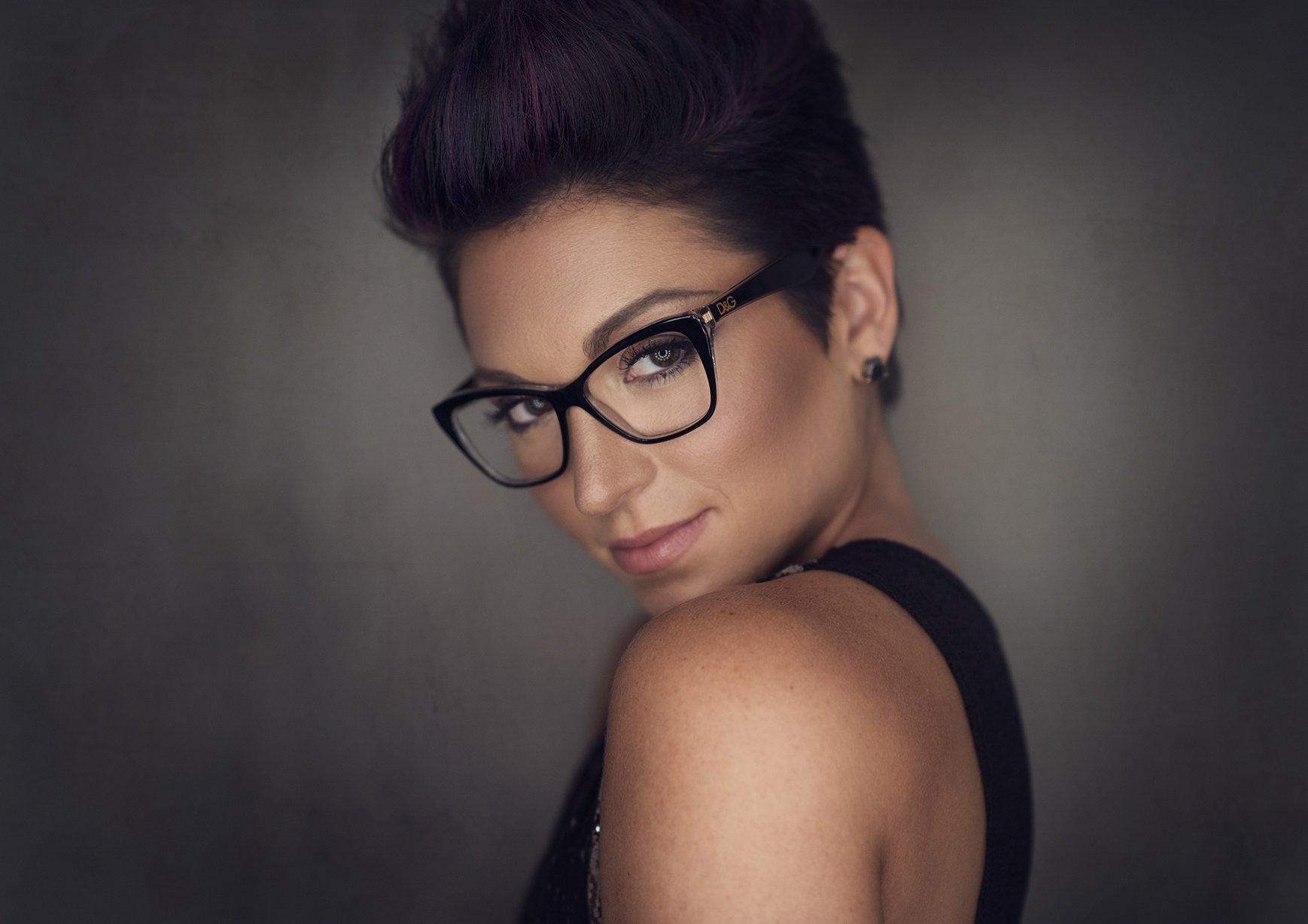 HD PC desktop wallpaper of a female model with short dark hair and glasses, gazing over her shoulder against a soft gray background.