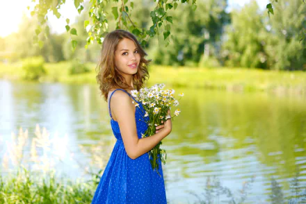 A smiling brunette woman with brown eyes wearing a blue dress holds a bouquet of flowers while standing outdoors by a serene lake. The image is a HD desktop wallpaper and background.
