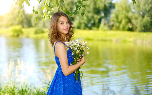 A smiling brunette woman with brown eyes wearing a blue dress holds a bouquet of flowers while standing outdoors by a serene lake. The image is a HD desktop wallpaper and background.
