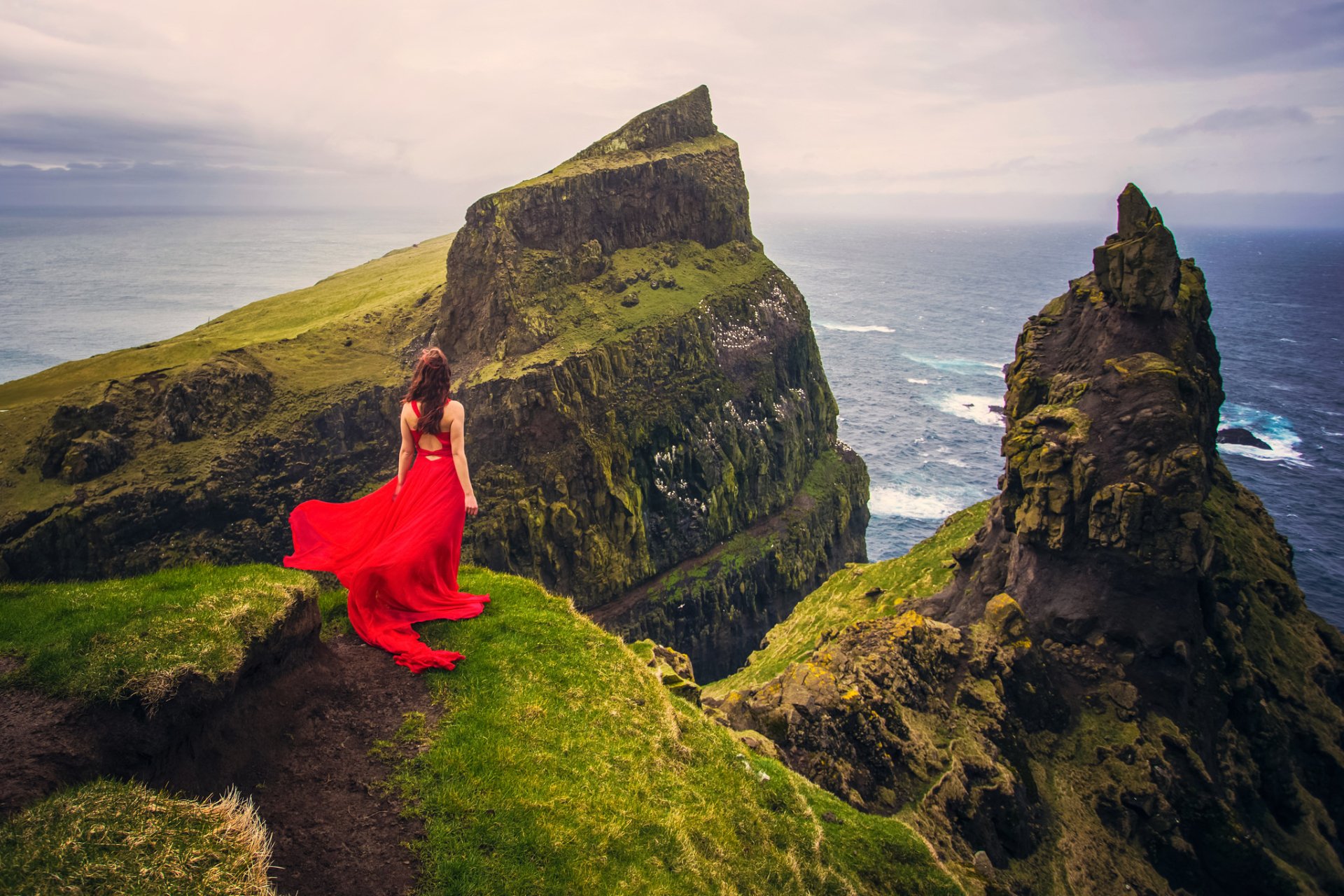 A woman in a red dress stands on a cliff, overlooking the vast sea and rugged stone mountains under a cloudy horizon. The image is a HD desktop wallpaper and background.