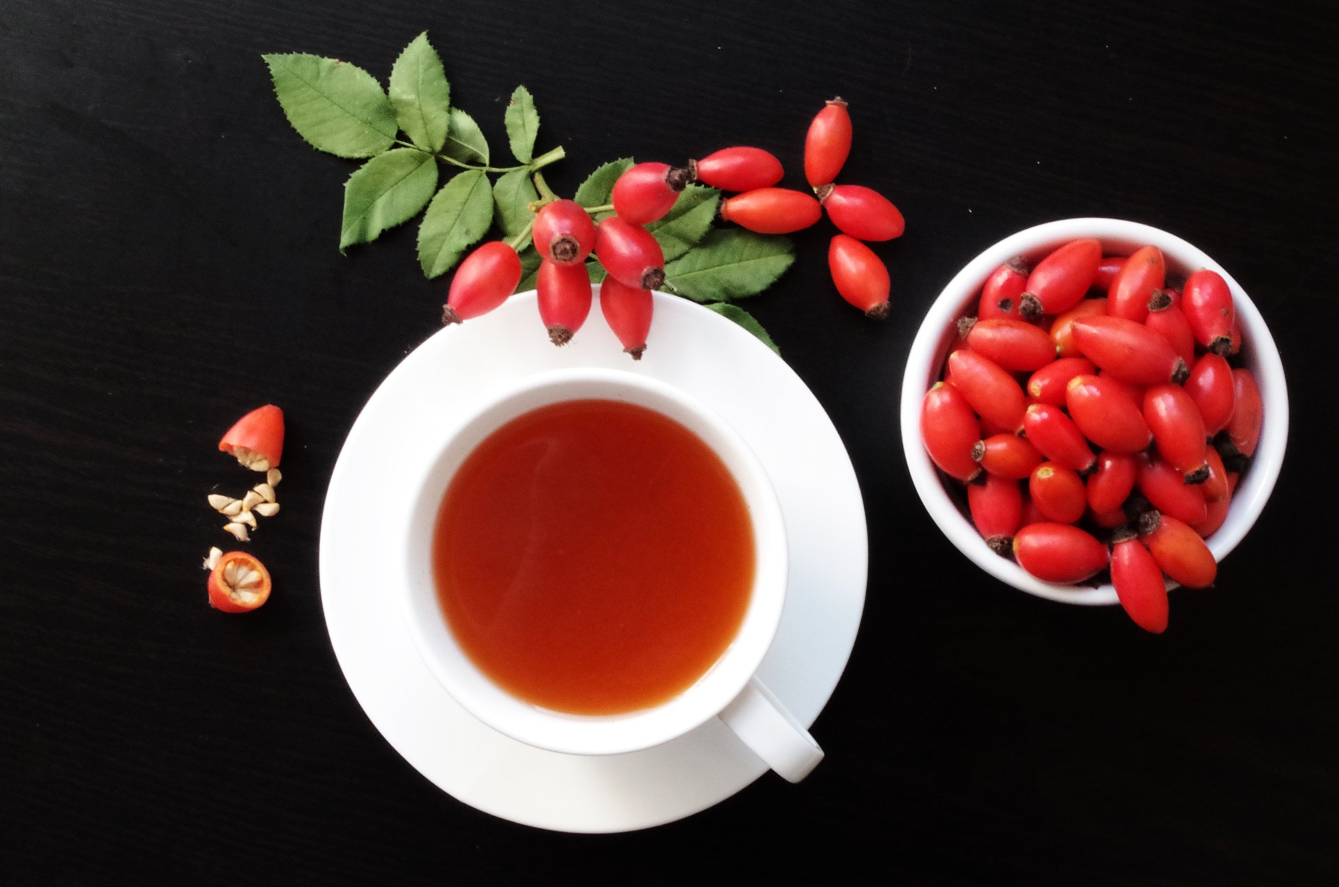 A 4K Ultra HD PC wallpaper featuring a cup of tea surrounded by fresh dog rose berries and a bowl of vibrant red dog rose fruits on a black background.