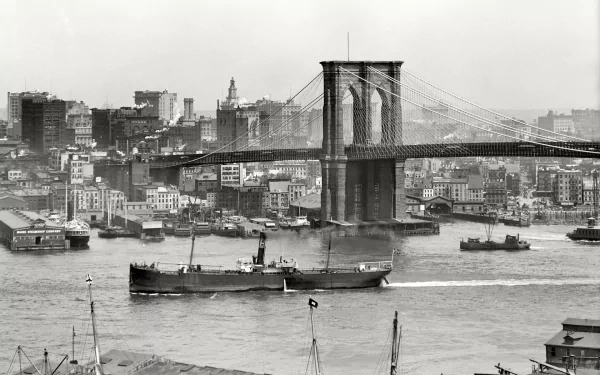 Black and white HD desktop wallpaper of the Brooklyn Bridge spanning the river with boats and city buildings in New York, USA.