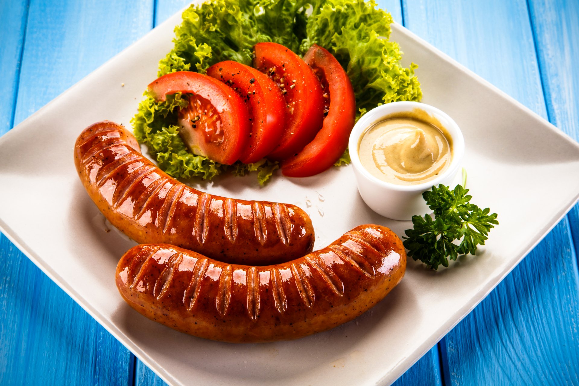 5K Ultra HD PC desktop wallpaper: two grilled sausages on a white plate with lettuce, sliced tomato, dipping sauce and parsley on a blue wood background.