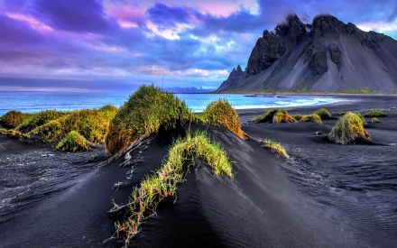 Vestrahorn Mountain rises dramatically above black sand dunes and ocean waves under a vibrant sky in Iceland, showcasing stunning natural beauty.