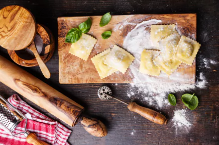 5K Ultra HD PC desktop wallpaper still life food scene: fresh ravioli dusted with flour on a wooden board, basil leaves, rolling pin and pastry cutter on a dark kitchen tabletop.