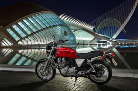 Red Honda CB1100 motorcycle parked at night beside an illuminated modern building reflected in water — 4K Ultra HD PC desktop wallpaper/background.