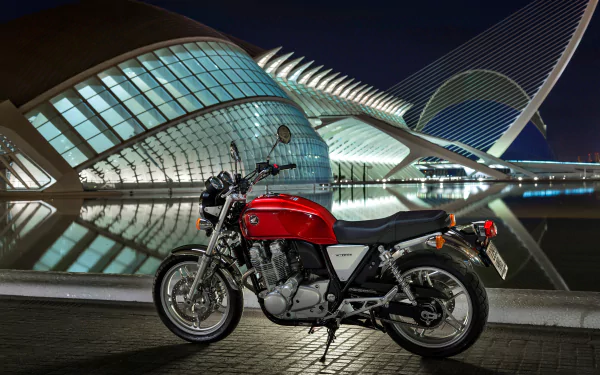 Red Honda CB1100 motorcycle parked at night beside an illuminated modern building reflected in water — 4K Ultra HD PC desktop wallpaper/background.