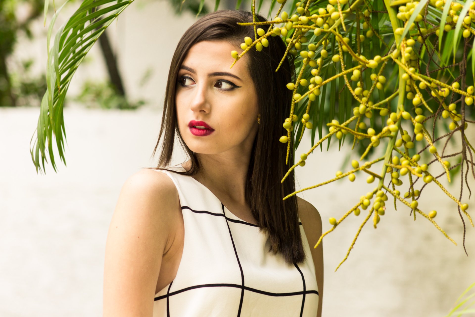 A brunette woman with brown eyes and red lipstick poses outdoors, surrounded by greenery. This HD desktop wallpaper features a stylish model in a white and black checkered dress.