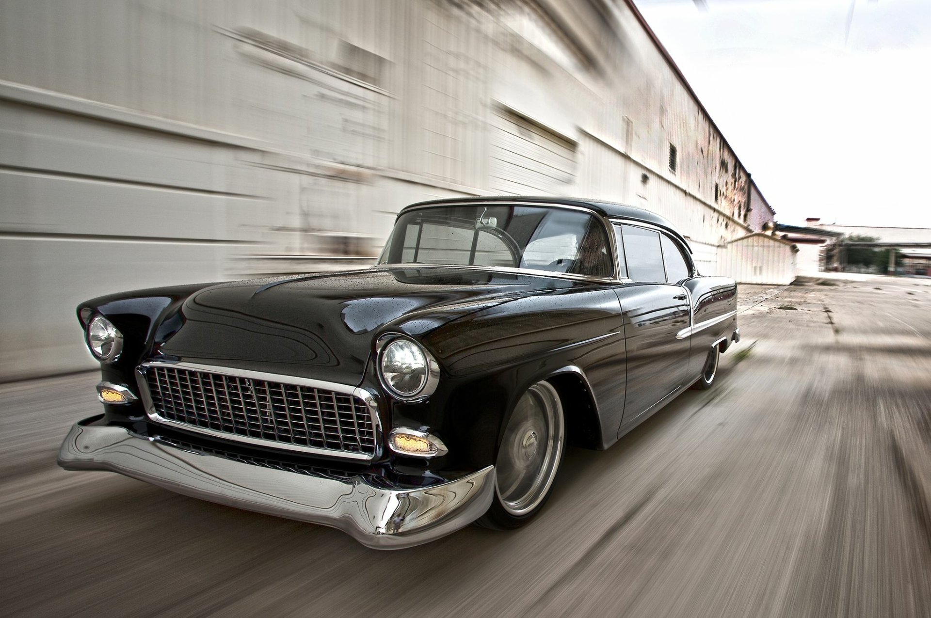 Black hot rod 1955 Chevrolet Bel Air vehicle speeding past industrial buildings, HD PC desktop wallpaper background.