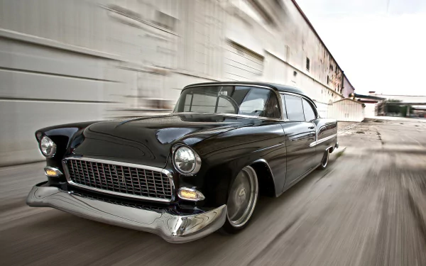 Black hot rod 1955 Chevrolet Bel Air vehicle speeding past industrial buildings, HD PC desktop wallpaper background.