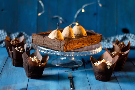 HD desktop wallpaper featuring a still life of a rich chocolate dessert topped with whole pears, surrounded by chocolate cupcakes on a blue wooden surface.