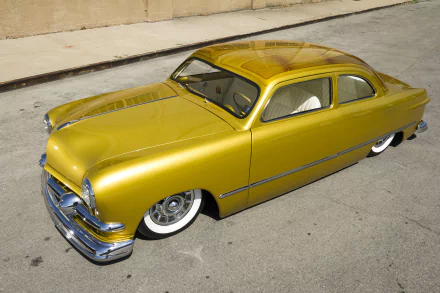 HD wallpaper featuring a golden 1951 Ford Custom lowrider hot rod with sleek lines and whitewall tires, parked on a sunlit urban street.