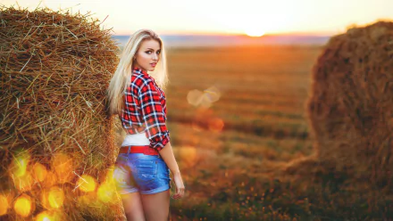 A blonde woman in shorts and a plaid shirt stands next to a haystack in a sunlit field, creating a picturesque scene. This HD desktop wallpaper and background exudes a warm, rustic charm.