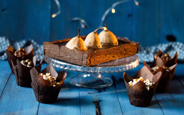HD desktop wallpaper featuring a still life of a rich chocolate dessert topped with whole pears, surrounded by chocolate cupcakes on a blue wooden surface.