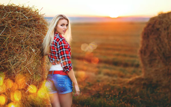 A blonde woman in shorts and a plaid shirt stands next to a haystack in a sunlit field, creating a picturesque scene. This HD desktop wallpaper and background exudes a warm, rustic charm.