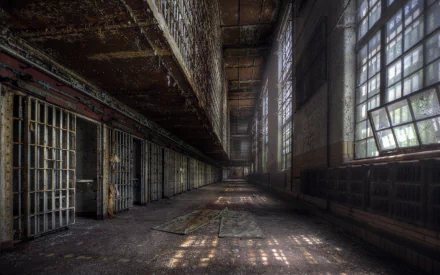 A haunting view of an abandoned prison corridor, featuring rusted bars and dust-covered floors, illuminated by sunlight filtering through tall, grimy windows.