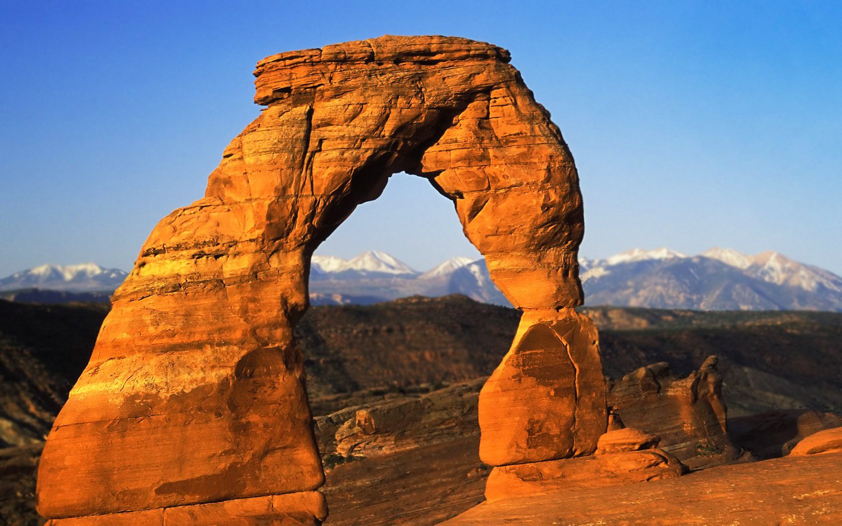 A stunning desert landscape featuring a towering rock arch against a clear blue sky, with distant mountains visible in the background. This HD wallpaper captures the essence of nature's beauty.