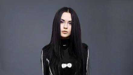 A brunette woman models a sleek latex outfit, showcasing her striking look and piercing features against a minimalist gray background. High-definition desktop wallpaper.