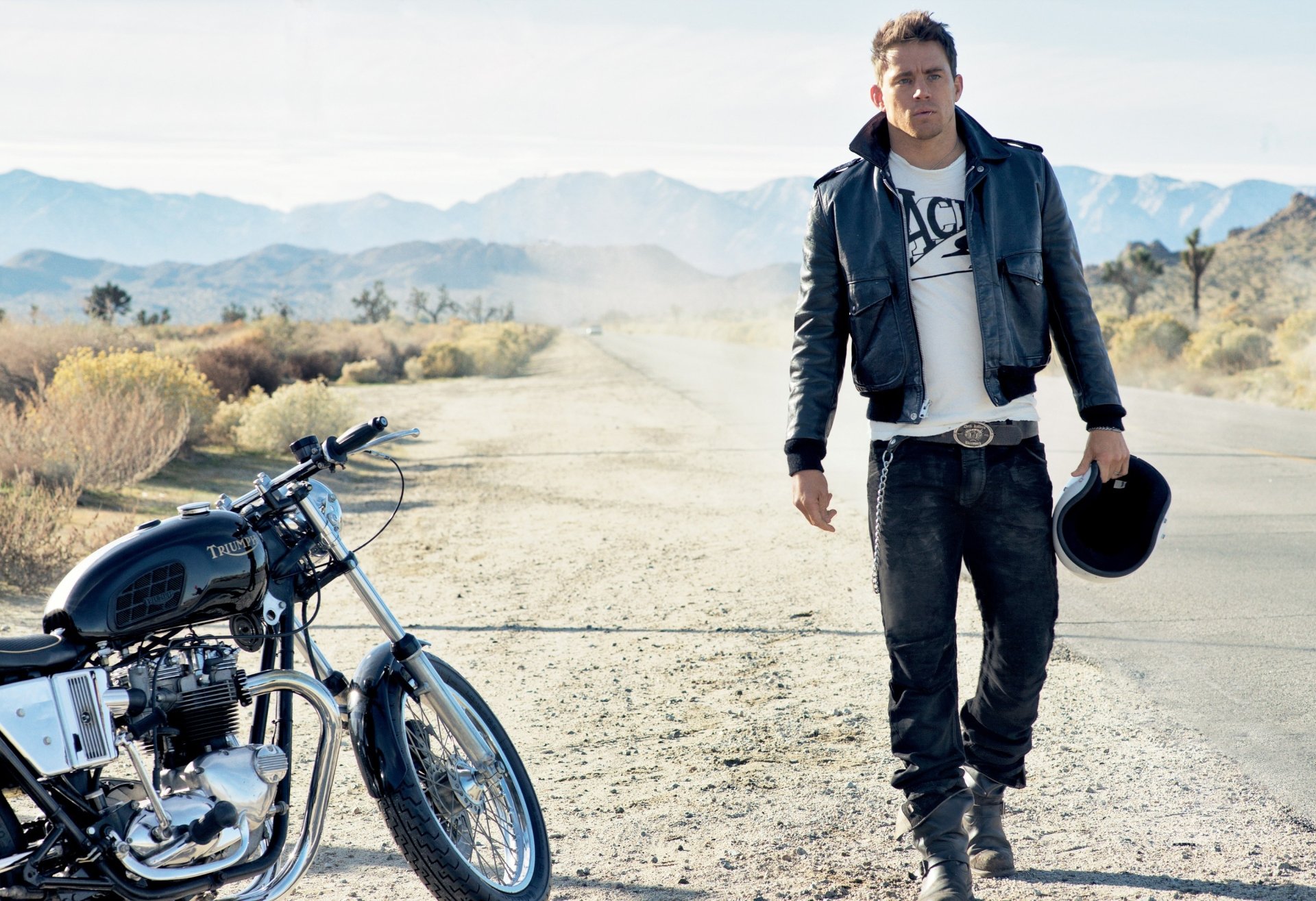 2K Quad HD PC wallpaper: American actor in a leather jacket walking along a desert road beside a Triumph motorcycle, holding a helmet.