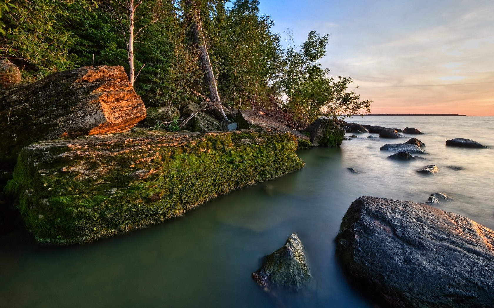 HD PC wallpaper showcasing a serene coastline with moss-covered rocks, calm water, and lush green trees under a soft evening sky.
