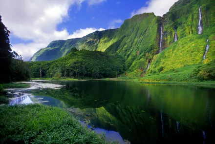 A stunning Azores landscape showcasing a lush green forest, towering mountains, and a serene waterfall cascading into a tranquil lake, reflecting the vibrant nature.