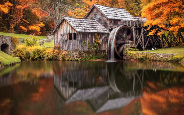 HD desktop wallpaper showing a rustic man-made watermill surrounded by vibrant fall foliage, reflected clearly in the calm water below.